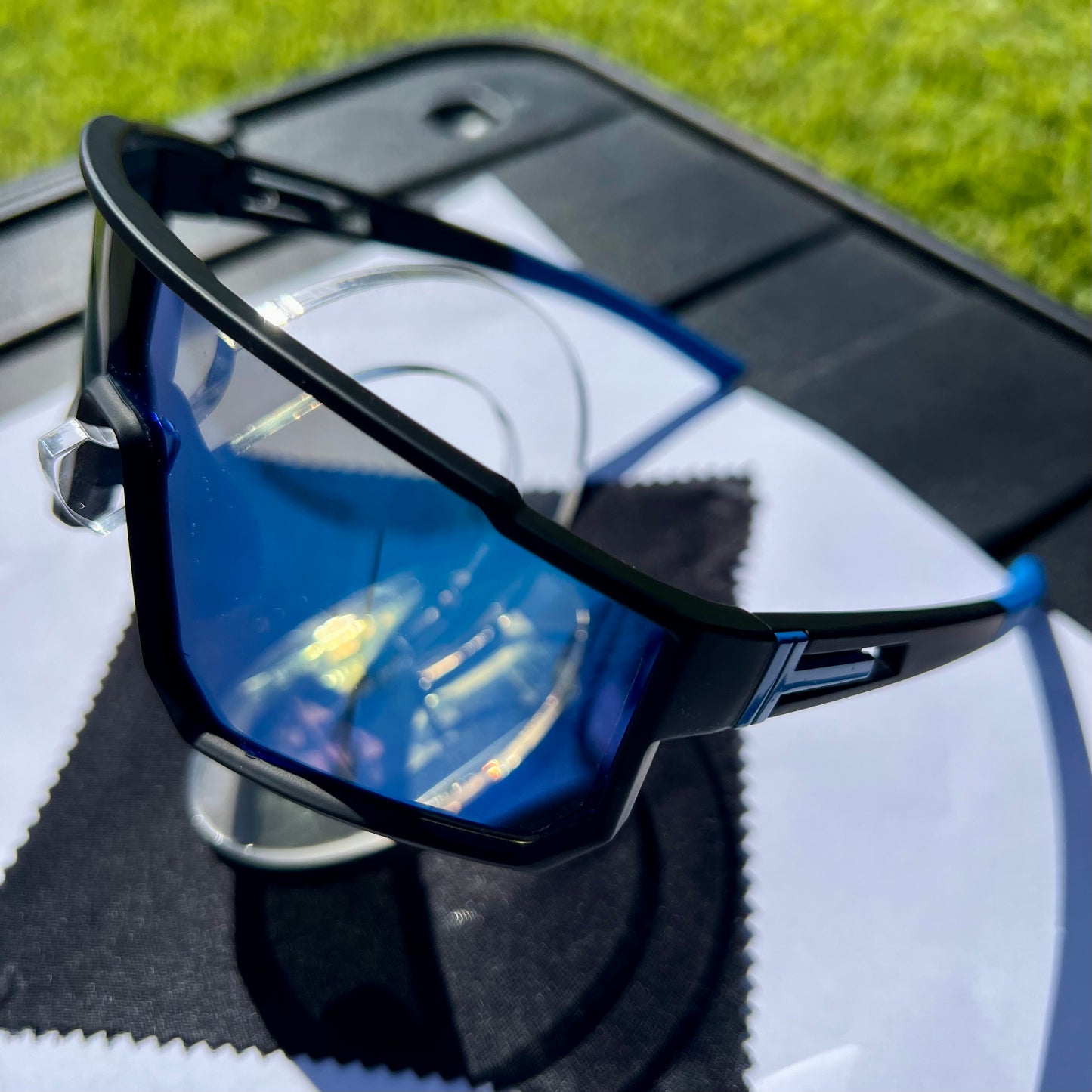 A pair of Sportiq VividShield sunglasses with blue reflective lenses and a black frame, polarized UV400, polycarbonate frame with TAC lens, displayed on a grass background.
