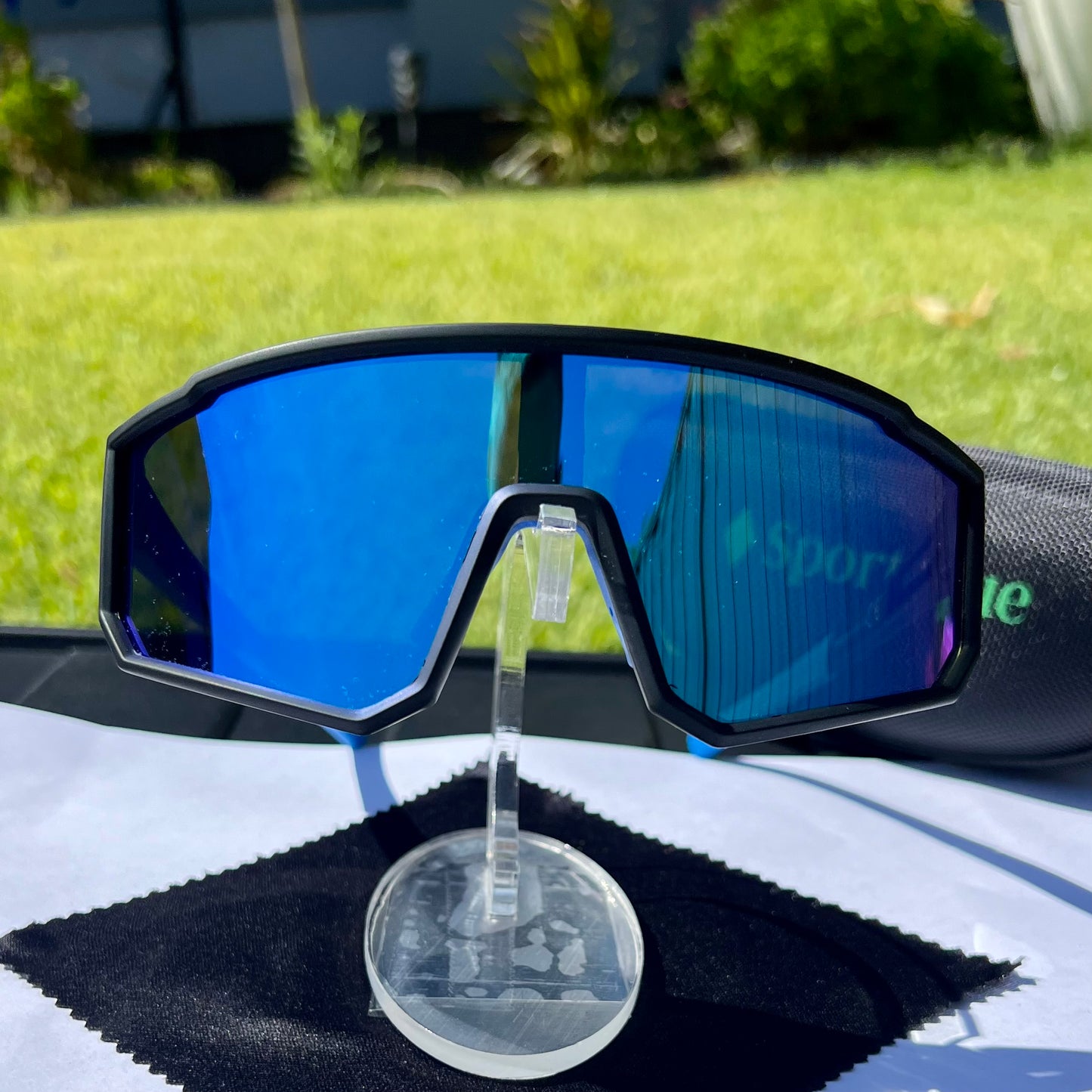 A pair of Sportiq VividShield sunglasses with blue reflective lenses and a black frame, polarized UV400, polycarbonate frame with TAC lens, displayed on a grass background.