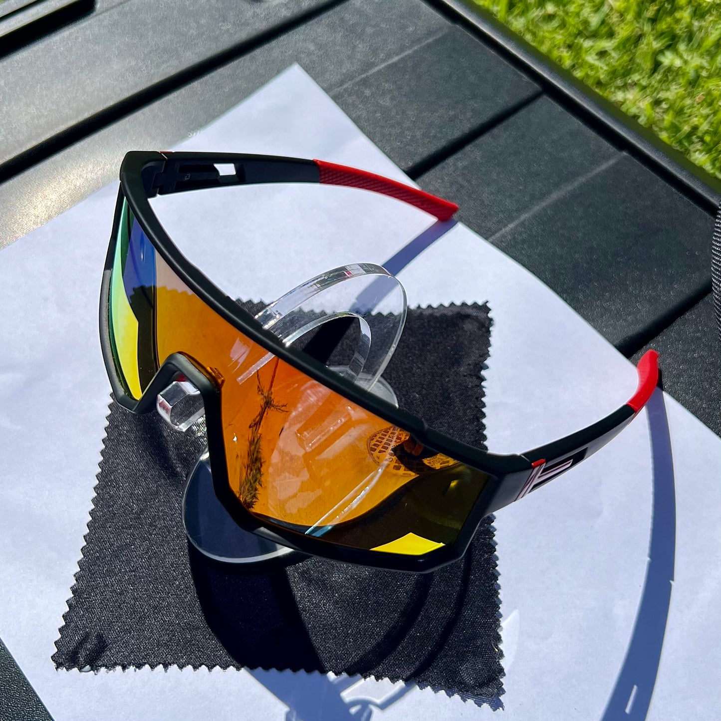 A pair of Sportiq VividShield sunglasses with black and red frame and red reflective lens, polarized UV400, polycarbonate frame with TAC lens, displayed on a grass background.