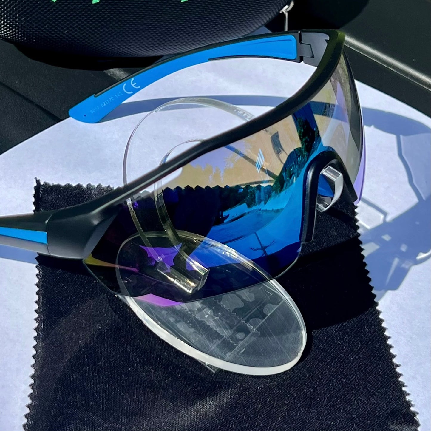 A pair of Sportiq ClarityX - Azure professional sport sunglasses with blue reflective lenses and black frame, polarized UV400, polycarbonate frame with TAC lens, displayed on a grass background.