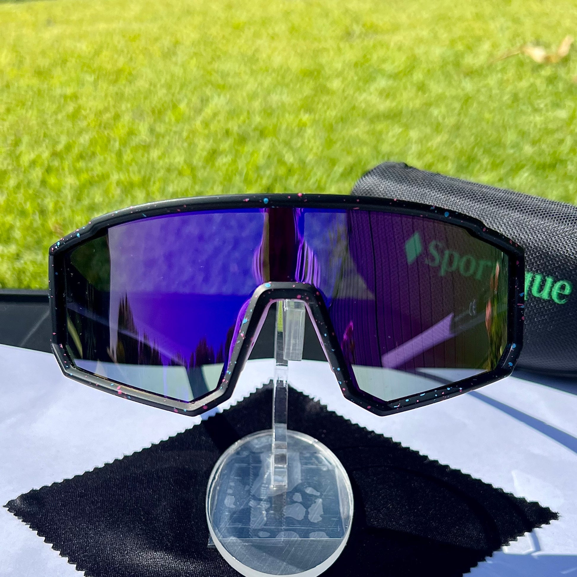 A pair of Sportiq VividShield sunglasses with a purple reflective lens and black frame,  polarized UV400, polycarbonate frame with TAC lens, displayed on a grass background.